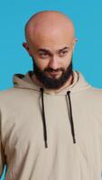 Vertical Video Middle eastern man acting casual on camera and showing groovy dance moves, feeling relaxed and carefree with beige hoodie. Young adult dancer having fun over blue background in studio. Handheld shot.