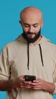 Vertical Video Arab person typing text messages on smartphone app, using mobile phone internet to check social media. Middle eastern guy browsing online websites in studio, blue backdrop. Camera 1.