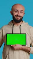 Vertical Video Arab guy holding device with greenscreen blank display, presenting isolated chromakey layout on tablet. Middle eastern adult using gadget to create promotional ad with copyspace. Camera 1.