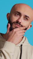 Vertical Video Thoughtful person being puzzled about a decision, standing over blue background in studio and thinking. Pensive middle eastern man being uncertain and doubtful about something. Camera 2.