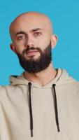 Vertical Video Young musician posing as a choir conductor in studio, conducting tunes for a band at classical show. Middle eastern person acting as bandmaster, serious expression over blue background. Camera 2.