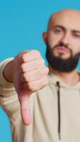 Vertical Video Middle eastern man raises palm and thumbs down in studio, expressing his rejection with stop symbol over blue background. Negative person showing denial and restriction. Camera 2. Handheld shot.