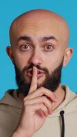 Vertical Video Middle eastern man doing hush silence symbol in studio, asking to keep a secret over blue background. Arab person creating mute gesture to be quiet in front of camera. Camera 2.