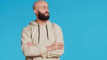 Young adult in a hoodie waiting for something in studio, standing with arms crossed over blue background. Middle eastern man being impatient on camera, punctual tensed person. Camera 2. video