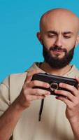 Vertikale Video froh Muslim Mann spielen Handy, Mobiltelefon Video Spiele im Studio, genießen online Wettbewerb mit Smartphone App. Arabisch Spieler haben Spaß mit Rollenspiel Herausforderung Wettbewerb. Kamera 2. Handheld Schuss.