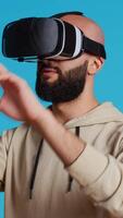 Vertical Video Middle eastern adult using virtual reality glasses in studio, having fun with metaverse artificial intelligence on vr headset. Young person posing over blue background, modern goggles. Camera 2.