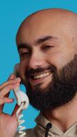 vertikal video mitten östra man talande till människor på fast telefon telefon ringa upp, svar retro maskin över blå bakgrund i studio. arab person njuter diskussion med kabel- telefon. kamera 2.