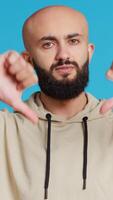 Vertical Video Middle eastern guy showing thumbs down sign on camera, giving dislike symbol and presenting his disappointment. Arab person feeling displeased with negative gesture in studio. Camera 2.