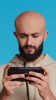 Vertical Video Middle eastern person playing mobile video games on app, standing over blue background and enjoying online tournament using mobile phone. Man having fun with gaming contest. Camera 2.