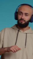 Vertical Video Muslim person listening to happy tunes on headphones, having fun with radio songs to feel relaxed and happy. Middle eastern guy dancing with headset over blue background. Camera 2.