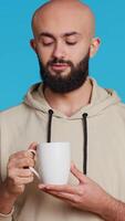 Vertical Video Muslim person enjoying fresh caffeine aroma in studio, drinking cup of coffee and standing over blue background. Middle eastern adult smelling beverage and taking a sip. Camera 2. Handheld shot.