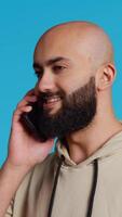 Vertical Video Middle eastern guy chatting with friends on phone call, answering smartphone while standing over blue background. Modern man discussing something on a remote network. Camera 2. Handheld shot.