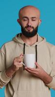 Vertical Video Muslim person enjoying fresh caffeine aroma in studio, drinking cup of coffee and standing over blue background. Middle eastern adult smelling beverage and taking a sip. Camera 1.