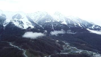 Top view of resort near mountains with snowy peaks. Small ski resort at foot of mountain with snowy peaks in fog in cloudy weather video