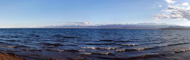 Blue calm water in Issyk-Kul lake with mountains on background at summer day - wide panorama photo