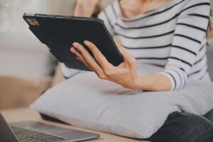 joven atractivo asiático mujer descansando utilizando hojeada tableta computadora en sofá a hogar, contento niña sentado en sofá relajarse leyendo digital artilugio con entusiasmado, comunicación y estilo de vida concepto. foto