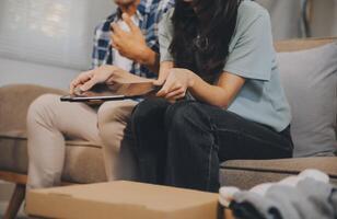 Two Asian blogger showing clothes in front of the camera to recording vlog video live streaming at her shop. Online Shopping Cart notion. on sofa at home photo