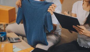 Two Asian blogger showing clothes in front of the camera to recording vlog video live streaming at her shop. Online Shopping Cart notion. on sofa at home photo
