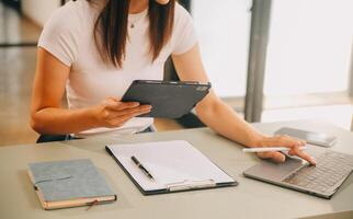 asiático mujer de negocios trabajando en ordenador portátil computadora Mira para trabajo en línea, Lanza libre mirando y mecanografía en cuaderno en mesa, estilo de vida de mujer estudiando en línea foto