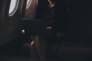 rubia hembra turista comprobación entrante notificación en teléfono inteligente sentado en asiento de avión con netbook.joven mujer de negocios compartir medios de comunicación desde teléfono en ordenador portátil computadora durante avión vuelo foto