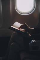 Blonde female tourist checking incoming notification on smartphone sitting on seat of airplane with netbook.Young businesswoman share media from telephone on laptop computer during plane flight photo