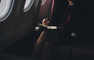 Blonde female tourist checking incoming notification on smartphone sitting on seat of airplane with netbook.Young businesswoman share media from telephone on laptop computer during plane flight photo