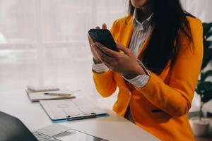 negocio mujer utilizando móvil teléfono durante trabajando en ordenador portátil computadora, surf el Internet, buscando negocio datos a moderno oficina. asiático mujer de negocios en línea trabajando en computadora a lugar de trabajo foto