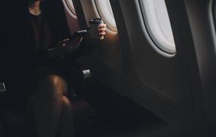 Blonde female tourist checking incoming notification on smartphone sitting on seat of airplane with netbook.Young businesswoman share media from telephone on laptop computer during plane flight photo