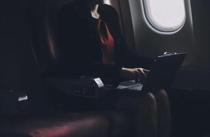 Blonde female tourist checking incoming notification on smartphone sitting on seat of airplane with netbook.Young businesswoman share media from telephone on laptop computer during plane flight photo