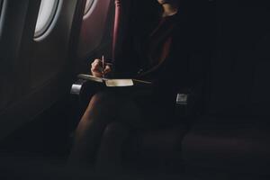 Blonde female tourist checking incoming notification on smartphone sitting on seat of airplane with netbook.Young businesswoman share media from telephone on laptop computer during plane flight photo