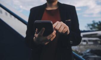 joven asiático mujer en internacional aeropuerto, utilizando móvil teléfono inteligente y comprobación vuelo a el vuelo información tablero foto