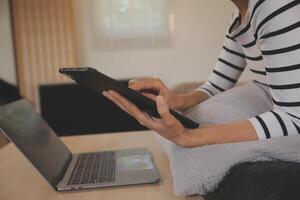 joven atractivo asiático mujer descansando utilizando hojeada tableta computadora en sofá a hogar, contento niña sentado en sofá relajarse leyendo digital artilugio con entusiasmado, comunicación y estilo de vida concepto. foto