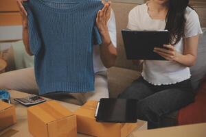 Two Asian blogger showing clothes in front of the camera to recording vlog video live streaming at her shop. Online Shopping Cart notion. on sofa at home photo