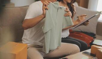 Two Asian blogger showing clothes in front of the camera to recording vlog video live streaming at her shop. Online Shopping Cart notion. on sofa at home photo