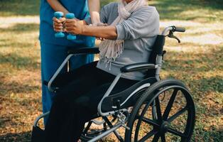 Young nurse or physiotherapist in scrubs helping a happy retired old woman do fitness exercises with light weight dumbbells at home. Concept of physiotherapy for seniors photo