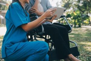 Elderly asian senior woman on wheelchair with Asian careful caregiver and encourage patient, walking in garden. with care from a caregiver and senior health insurance in sun light photo