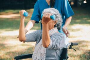 Young nurse or physiotherapist in scrubs helping a happy retired old woman do fitness exercises with light weight dumbbells at home. Concept of physiotherapy for seniors photo