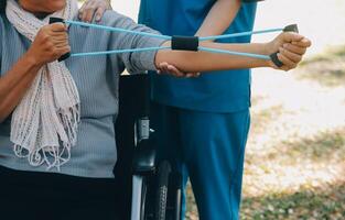 Young nurse or physiotherapist in scrubs helping a happy retired old woman do fitness exercises with light weight dumbbells at home. Concept of physiotherapy for seniors photo