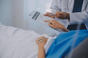 The senior woman specialist doctor with stethoscope checking up happy young Asian female patient lying in bed with receiving an intravenous saline drip in recovery room. Healthcare, medical insurance. photo