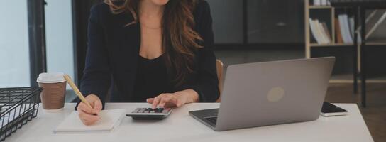 Asian Businesswoman Using laptop computer and working at office with calculator document on desk, doing planning analyzing the financial report, business plan investment, finance analysis concept. photo