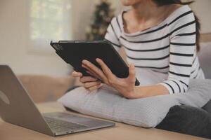 joven atractivo asiático mujer descansando utilizando hojeada tableta computadora en sofá a hogar, contento niña sentado en sofá relajarse leyendo digital artilugio con entusiasmado, comunicación y estilo de vida concepto. foto