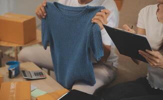 Two Asian blogger showing clothes in front of the camera to recording vlog video live streaming at her shop. Online Shopping Cart notion. on sofa at home photo