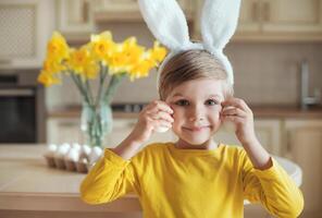 Closeup portrait of cute child in rabbit ears holding eggs for easter holiday on the kitchen. Bouquet of yellow daffodils flowers on background. photo