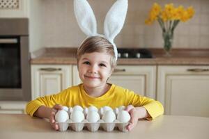 linda sonriente chico en amarillo ropa y Conejo orejas preparando para Pascua de Resurrección, participación un paquete de blanco huevos en el mesa en el cocina foto