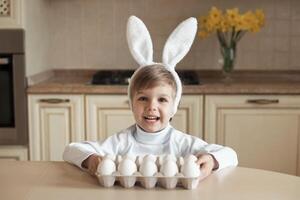 Lifestyle portrait of laughing little child celebrating easter at home kithen, wearing bunny ears, holding chiken no colored white eggs. Eco easter concept. photo