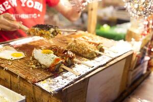 okonomiyaki con brochetas parrilla cocido en el tostador a noche local mercado foto