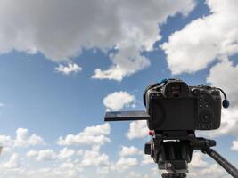 black professional digital camera with flip screen on a tripod pointed at blue sky with white clouds photo