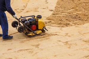 Worker using vibratory plate compactor for compaction sand during path construction. photo