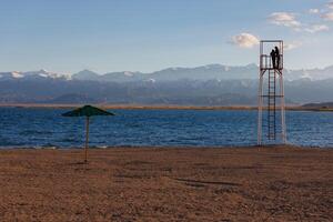 two person silhouettes on observation tower near steel umbrella on beach of mountain lake at sunny evening photo