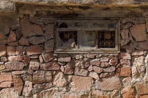 small window with broken glass and metal grate in ancient wall at sunny day, closeup full-frame background photo